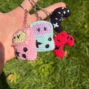 May include: Four crocheted keyrings in the shape of game controllers. The keyrings are in pink, light green, light blue, black, and red. Each keyring has a silver chain and ring. The keyrings are handmade.