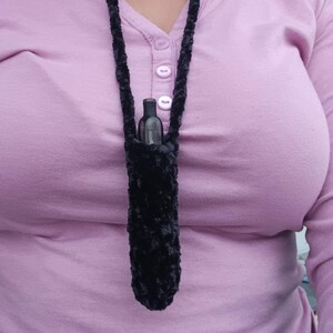 Może przedstawiać: Czarny, pluszowy, szydełkowy naszyjnik z pasującą torebką, w kt&oacute;rej znajduje się mała, ciemna butelka. Naszyjnik jest noszony na szyi, a torebka spoczywa na wysokości klatki piersiowej. Tło to jasnofioletowa koszula z długim rękawem.