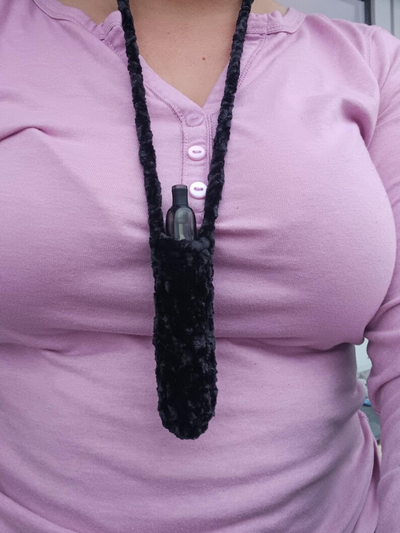 Może przedstawiać: Czarny, pluszowy, szydełkowy naszyjnik z pasującą torebką, w kt&oacute;rej znajduje się mała, ciemna butelka. Naszyjnik jest noszony na szyi, a torebka spoczywa na wysokości klatki piersiowej. Tło to jasnofioletowa koszula z długim rękawem.