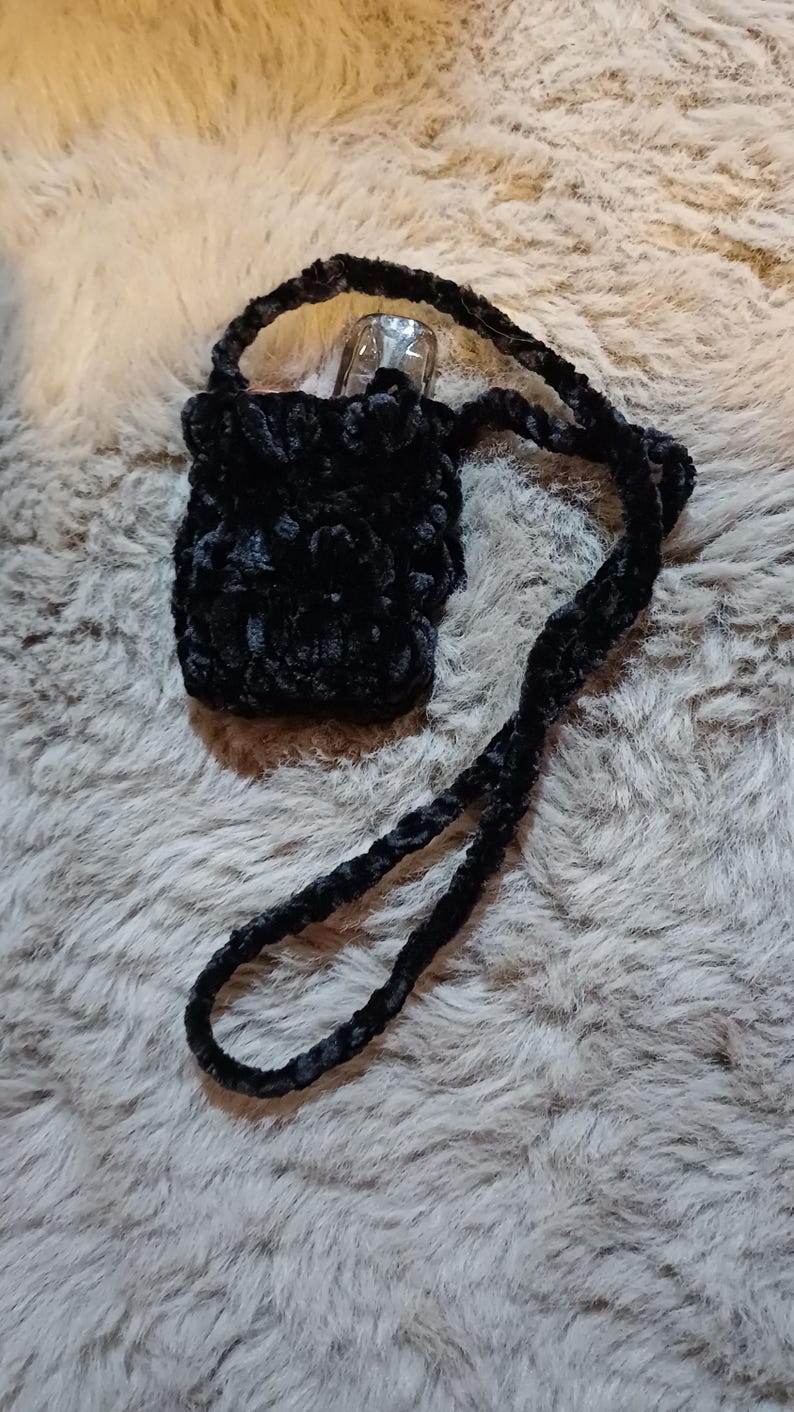 Może przedstawiać: Mała, czarna, pluszowa torba z długim paskiem. Torba wykonana jest z miękkiego, puszystego materiału, prawdopodobnie do przenoszenia małego przedmiotu. Pasek r&oacute;wnież wykonany jest z tego samego materiału.