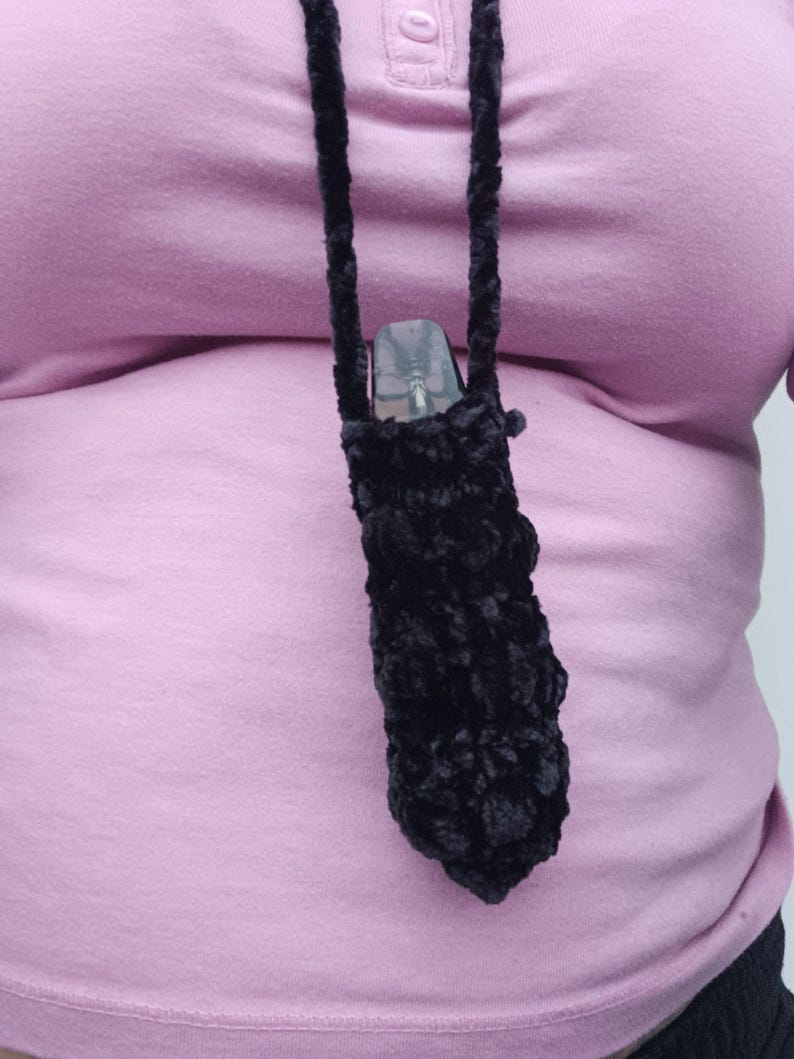 Może przedstawiać: Czarny, pluszowy, szydełkowy uchwyt na vape z pasującym sznurkiem, noszony na szyi. Uchwyt zawiera przezroczyste urządzenie vape. Tło to jasnor&oacute;żowa koszula.