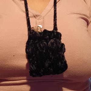 Może przedstawiać: Mała, czarna, pluszowa torebka z długim, pasującym paskiem. Torebka jest noszona na szyi i wydaje się być ręcznie robiona. Tło to jasnor&oacute;żowa koszula.