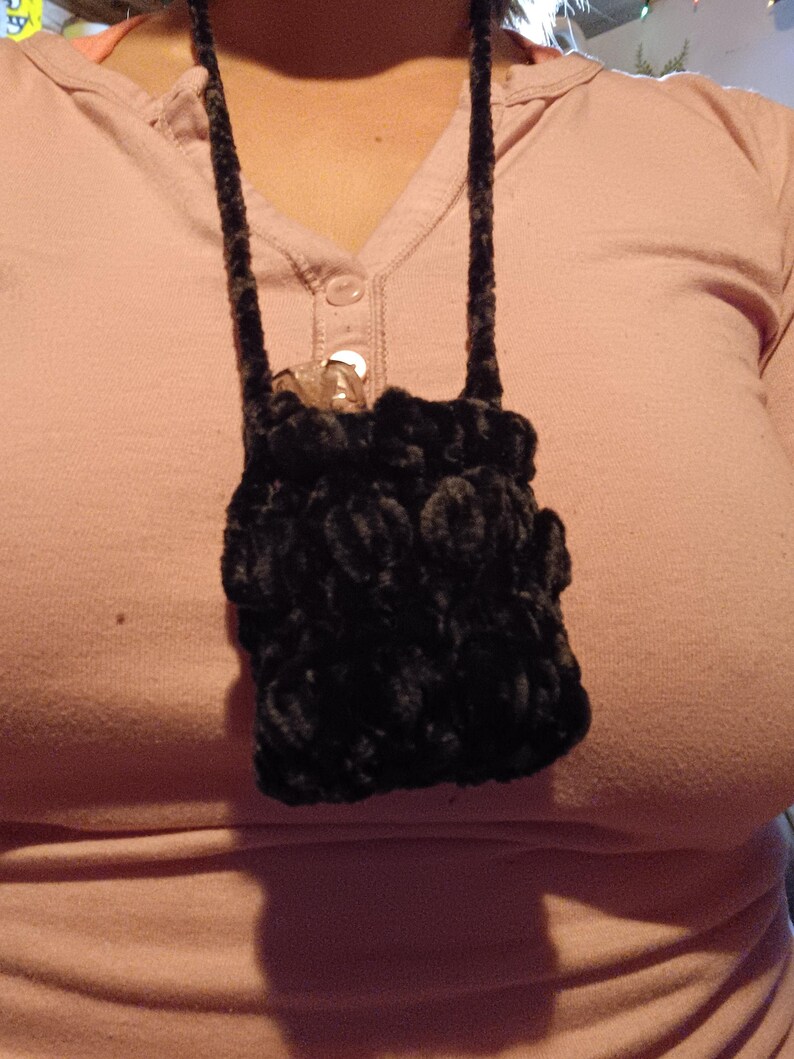 Może przedstawiać: Mała, czarna, pluszowa torebka z długim, pasującym paskiem. Torebka jest noszona na szyi i wydaje się być ręcznie robiona. Tło to jasnor&oacute;żowa koszula.