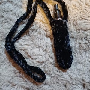 Może przedstawiać: Czarny, pluszowy, ręcznie robiony uchwyt na długopis vape z pasującym paskiem. Uchwyt jest przeznaczony do przenoszenia długopisu vape i jest pokazany na szarej, puszystej powierzchni. Pasek jest wystarczająco długi, aby można go było nosić na szyi.