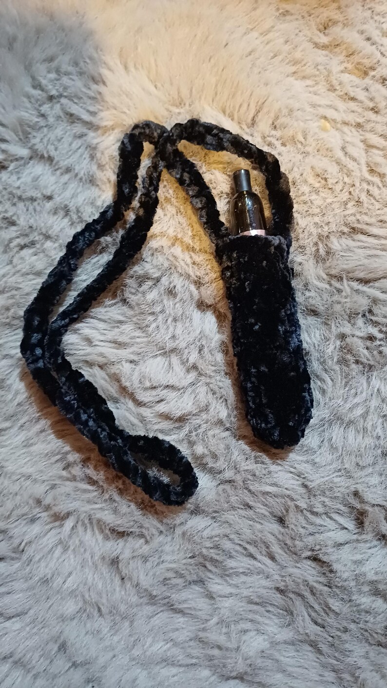 Może przedstawiać: Czarny, pluszowy, ręcznie robiony uchwyt na długopis vape z pasującym paskiem. Uchwyt jest przeznaczony do przenoszenia długopisu vape i jest pokazany na szarej, puszystej powierzchni. Pasek jest wystarczająco długi, aby można go było nosić na szyi.