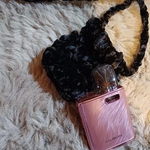 Może przedstawiać: R&oacute;żowe urządzenie do waporyzacji z napisem "CALIBURN", spoczywające w czarnej, puszystej, szydełkowej torebce z pasującym paskiem. Przedmioty znajdują się na kudłatej, jasnej powierzchni.