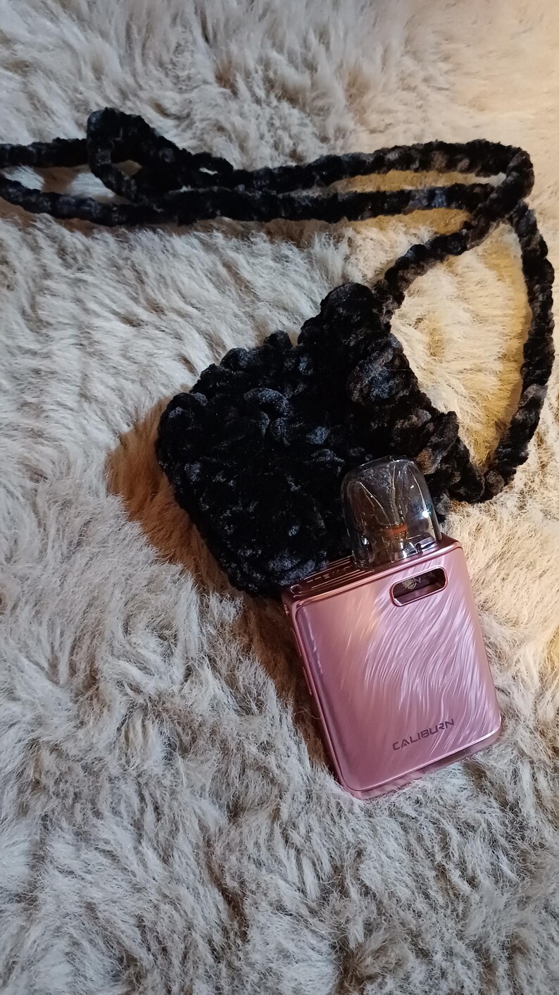 Może przedstawiać: R&oacute;żowe urządzenie do waporyzacji z napisem "CALIBURN", spoczywające w czarnej, puszystej, szydełkowej torebce z pasującym paskiem. Przedmioty znajdują się na kudłatej, jasnej powierzchni.