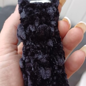 Może przedstawiać: Czarny, pluszowy pokrowiec na urządzenie do waporyzacji. Pokrowiec wykonany jest z miękkiego, puszystego materiału i zakrywa urządzenie, z przezroczystym ustnikiem widocznym na g&oacute;rze. Pokrowiec jest trzymany w dłoni.