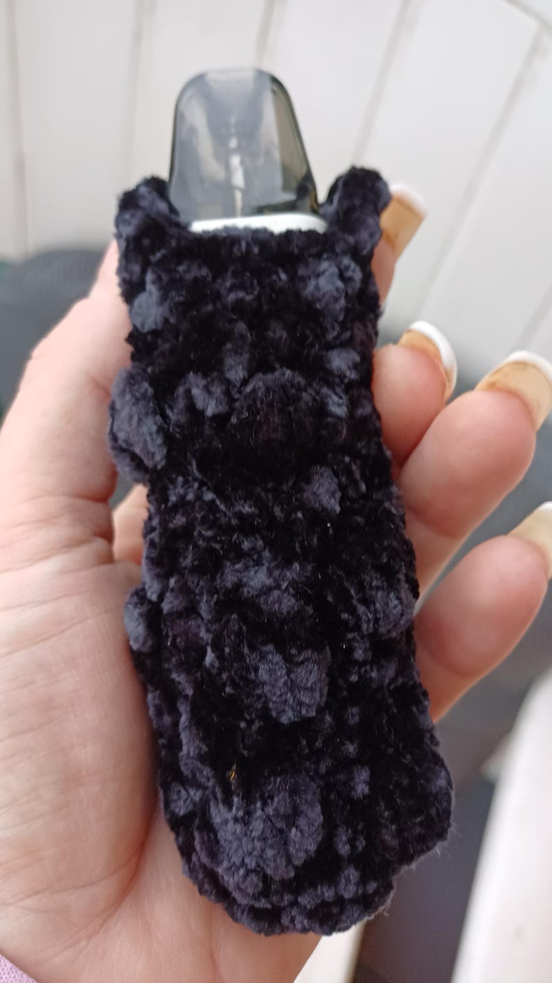 Może przedstawiać: Czarny, pluszowy pokrowiec na urządzenie do waporyzacji. Pokrowiec wykonany jest z miękkiego, puszystego materiału i zakrywa urządzenie, z przezroczystym ustnikiem widocznym na g&oacute;rze. Pokrowiec jest trzymany w dłoni.