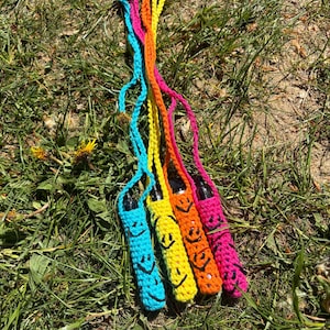 Op de afbeelding: Een verzameling van vijf kleurrijke gehaakte vape pen houders met smiley gezichten. De houders zijn in tinten blauw, geel, oranje en roze, elk met een bijpassend koord. De pennen liggen op een bed van groen gras.