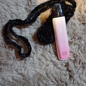 Może przedstawiać: R&oacute;żowe i białe urządzenie do waporyzacji z czarnym ustnikiem i literami "XK" wydrukowanymi z boku. Jest przymocowany do czarnej, pluszowej smyczy. Urządzenie spoczywa na teksturowanej, szarej powierzchni.