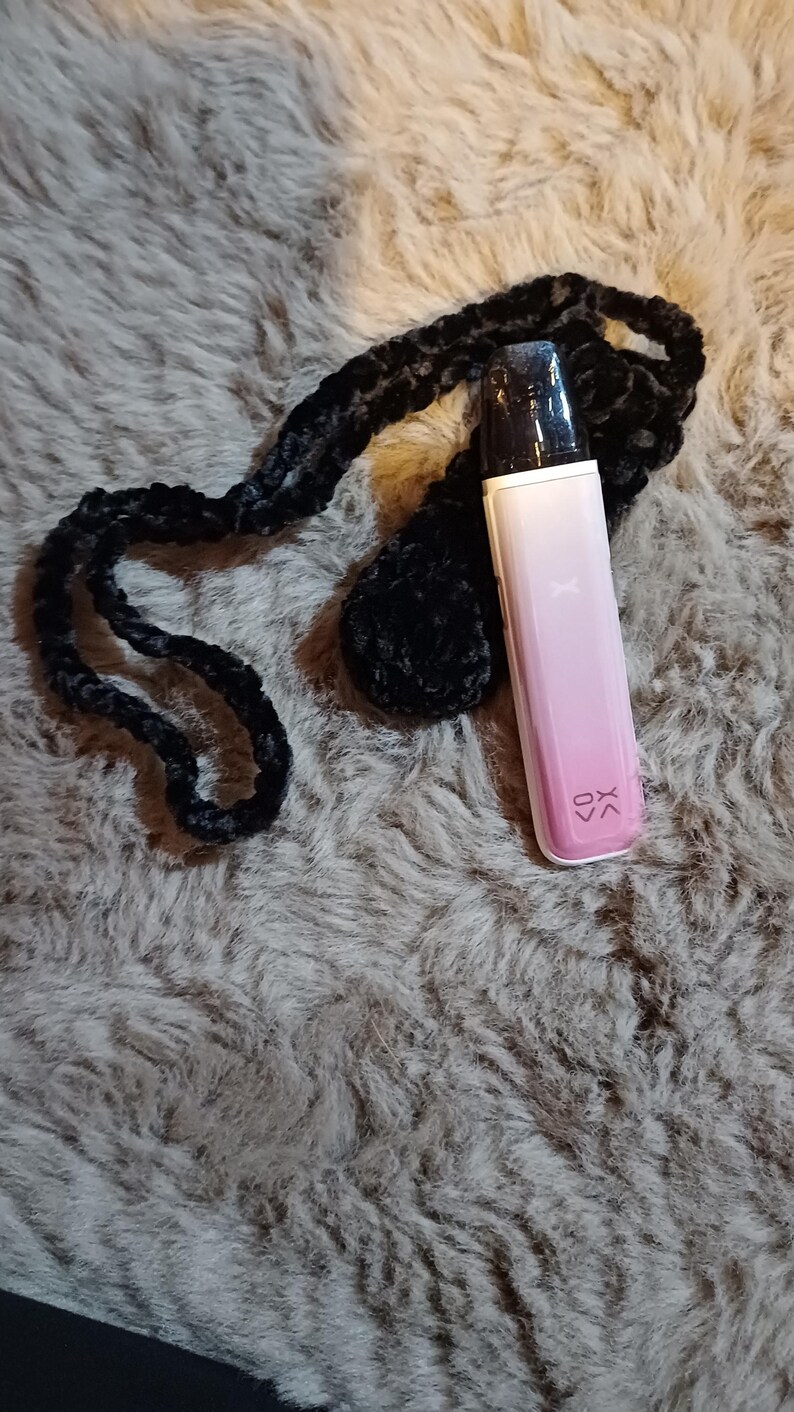 Może przedstawiać: R&oacute;żowe i białe urządzenie do waporyzacji z czarnym ustnikiem i literami "XK" wydrukowanymi z boku. Jest przymocowany do czarnej, pluszowej smyczy. Urządzenie spoczywa na teksturowanej, szarej powierzchni.