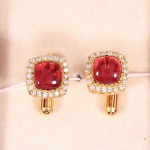 Puede incluir: Un par de gemelos dorados con piedras rojas cuadradas rodeadas por un halo de pequeñas piedras transparentes. Los gemelos se presentan en un joyero de terciopelo rojo con interior color crema.