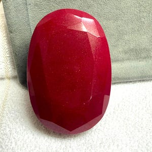 Könnte beinhalten: Ein großer, facettierter, oval geformter Rubin-Edelstein. Der Edelstein hat eine tiefe, satte rote Farbe mit einer polierten Oberfläche. Die Facetten erzeugen ein Lichtspiel auf der Oberfläche des Steins.
