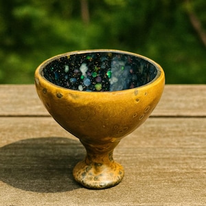May include: A handmade ceramic bowl with a pedestal base. The exterior is a warm, golden-yellow color, while the interior is a dark, speckled blue with hints of green and purple. The bowl is sitting on a wooden surface.