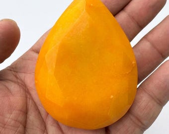 May include: A faceted, teardrop-shaped gemstone in a vibrant orange hue. The gem is held in an open hand, showcasing its polished surface and geometric cuts. The gemstone's color ranges from light to deep orange.