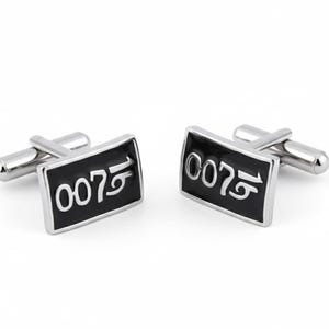Op de afbeelding: Een paar rechthoekige manchetknopen met een zwarte emaille voorkant en zilverkleurige metalen rand. De manchetknopen tonen het nummer "007" in wit. Gepresenteerd op een witte achtergrond.