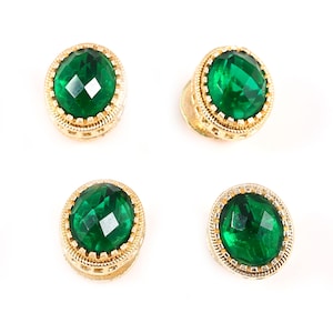 May include: Four emerald green oval-shaped gemstones with gold-colored, ornate settings. The gemstones have a faceted cut, and the settings feature a detailed, textured design. The items are displayed against a white background.