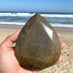 Op de afbeelding: Een grote, druppelvormige edelsteen met een facetgeslepen bovenkant. De steen heeft een mix van groene en bruine tinten, met een gepolijst oppervlak dat licht reflecteert. De achtergrond toont een strand en de oceaan.
