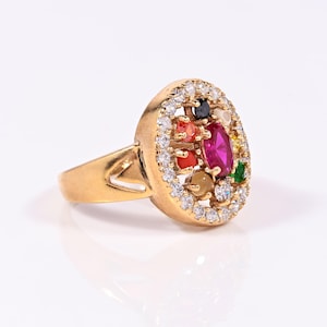 May include: A gold-colored ring featuring a cluster of colorful gemstones. The ring's centerpiece is a large, oval-shaped magenta stone, surrounded by smaller gems in various colors. The band is a simple, polished gold, and the ring is set against a white background.