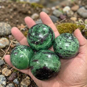 Op de afbeelding: Vier gepolijste groene en rode robijn-zoisiet bollen worden in een hand gehouden. De bollen hebben een gespikkelde uitstraling met donkergroene en zwarte vlekken en rode aderen. De achtergrond is wazig, met rotsen en groen.