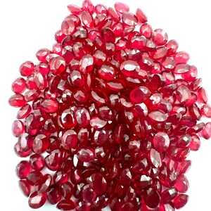 May include: A pile of faceted, oval-shaped, deep red gemstones. The gems are scattered across a white surface, catching the light and displaying a brilliant shine. The gemstones are likely rubies, perfect for jewelry making.