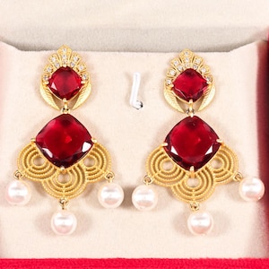 以下が含まれることがあります： 大きな正方形のルビーレッドの宝石が付いた、ゴールドトーンのシャンデリアイヤリングのペア。 イヤリングはフィリグリーデザインで、小さなクリアクリスタルと白いパールがアクセントになっています。 イヤリングは赤いジュエリーボックスに飾られています。