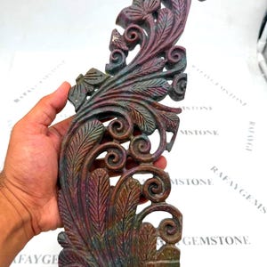Puede incluir: Una escultura de piedra preciosa tallada a mano con intrincados patrones de hojas y enredaderas en espiral. La piedra muestra una paleta de colores variados de rojo intenso, marrón terroso y tonos verdes apagados en su superficie.