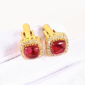 May include: A pair of gold cufflinks with square, ruby-red center stones. Each cufflink is surrounded by a halo of small, clear gemstones. The cufflinks are set against a white background.