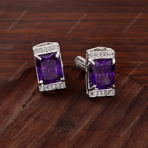 Puede incluir: Un par de gemelos rectangulares de plata, cada uno con una gran piedra preciosa de color púrpura intenso y corte esmeralda en el centro. Los bordes superior e inferior están acentuados con una hilera de pequeñas piedras claras y brillantes.