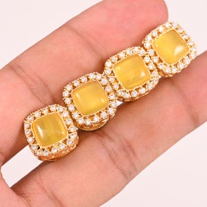 Peut inclure: Quatre bijoux carrés dorés, avec une pierre jaune centrale et une bordure de petits pierres précieuses transparentes. Les pièces sont disposées en rang, mettant en valeur leur design et leur savoir-faire.