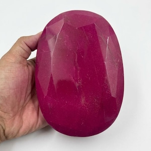 Pode incluir: Grande gema de rubi oval e facetada. O rubi é de uma cor vermelha profunda e rica, com múltiplas facetas que capturam a luz. A gema é segurada em uma mão contra um fundo branco.