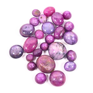 May include: A collection of polished gemstones in shades of purple and pink, with varying shapes and sizes. The stones range from small, round beads to larger, oval cabochons. The gemstones are arranged on a white background.