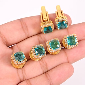 May include: Gold-toned cufflinks and stud earrings set. Each piece features a square, teal-colored gemstone surrounded by small, clear crystals. The cufflinks have a rectangular bar design, while the stud earrings have a post backing.