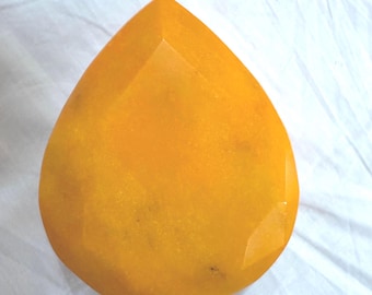 May include: A large, faceted, teardrop-shaped gemstone in a vibrant shade of yellow-orange. The gem has a polished surface with multiple facets, catching the light and creating a sparkling effect. The gemstone is set against a white background.