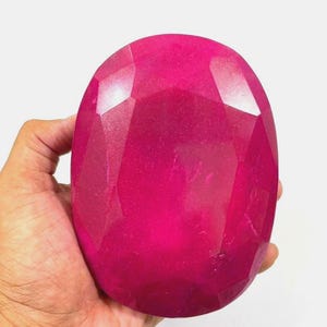 May include: A large, faceted, oval ruby gemstone. The gem is a vibrant pinkish-red, with multiple facets catching the light. The ruby is held in a hand against a white background, showcasing its size and colour.