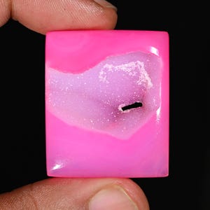 Puede incluir: Una gema rectangular y pulida de color rosa vibrante. La piedra presenta una zona cristalina más clara en el centro, con una inclusión lineal oscura. La gema se sostiene entre dos dedos sobre un fondo negro.