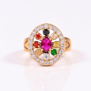 May include: Gold ring with a central oval pink gemstone, encircled by a halo of colourful gemstones and clear crystals. The ring has a decorative band with a textured design. The ring is set against a white background.