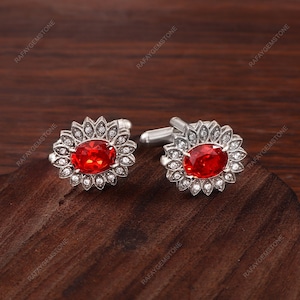 Puede incluir: Un par de gemelos plateados con una piedra preciosa roja de corte ovalado en el centro, rodeada por un halo de pequeñas piedras transparentes dispuestas en forma de pétalos. El engaste metálico pulido tiene un cierre de barra en T.