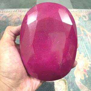 Könnte beinhalten: Ein großer, facettierter, oval geformter Edelstein in einem tiefen Magenta-Farbton. Der Edelstein wird in einer Hand gehalten, um seine Größe und polierten Oberflächen zu zeigen. Die Facetten fangen das Licht ein und erzeugen Glanzlichter.