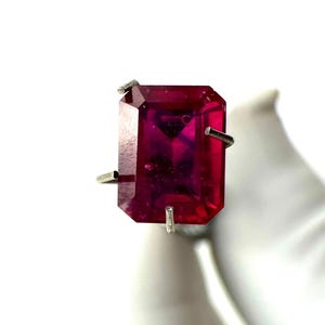May include: A striking ring featuring a large, rectangular, ruby-red gemstone. The gemstone is set in a silver-coloured metal band. The ring's design has a bold, geometric shape, with a polished, faceted surface.