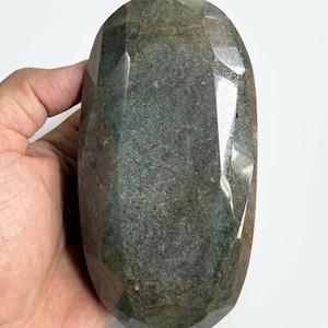 May include: A large, faceted gemstone with a dark green and brown colour. The stone has an oval shape and is held in a hand against a white background. The facets create a sparkling effect.