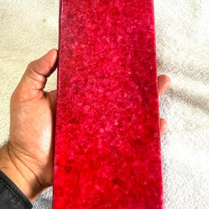 Könnte beinhalten: Ein rechteckiger, roter Dekorationsstein mit einem gesprenkelten Muster. Der Stein wird in einer Hand gehalten, wodurch seine polierte Oberfläche und seine leuchtende Farbe zur Geltung kommen. Die Abmessungen des Steins sind nicht sichtbar.