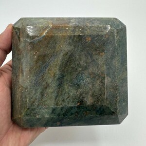 May include: A large, rectangular, gemstone-like object with beveled edges. The stone has a mottled appearance with shades of green, brown, and blue. The object is held in a hand, showcasing its size and texture. The overall aesthetic suggests a decorative or collectible item.