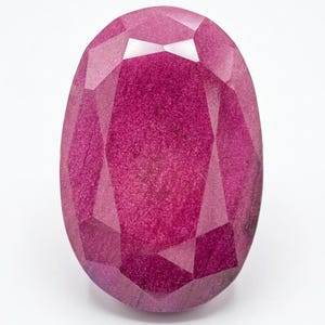 May include: A faceted, oval-shaped ruby gemstone. The gem is a deep, saturated pinkish-red color with multiple facets that reflect light. The gemstone is set against a white background.