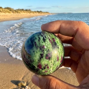 Könnte beinhalten: Ein grüner und roter kugelartiger Edelstein, gehalten in einer Hand, vor dem Hintergrund eines Sandstrandes und des Ozeans. Der Stein hat ein gesprenkeltes Muster aus Grün und Dunkelrot. Strand und Ozean sind im Hintergrund sichtbar.