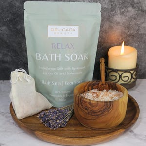 May include: A green and white bag of Himalayan bath salts with lavender, jojoba oil, and botanicals. The bag is labeled "Relax Bath Soak" and "Delicada Beauty". The bag is sitting on a wooden tray with a small bowl of bath salts, a wooden spoon, a small bag of lavender, and a lit candle.