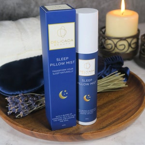 May include: Two blue bottles of sleep pillow mist with gold lettering and a crescent moon and stars design. The bottles are on a wooden tray with a blue silk eye mask and dried lavender.
