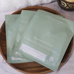May include: Three light green facial sheet mask packets with the text "DELICADA BEAUTY Nourish & Glow Moisturising Facial Sheet Mask" on a wooden tray. The packets have a white border and a graphic of a face. The tray is on a marble surface.