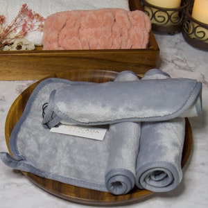 May include: Three gray microfiber makeup remover cloths rolled up and placed on a wooden plate. The cloths are labeled "Delicada Beauty".